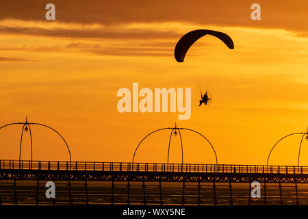 Ein motorbetriebener Handsegler, der in der Abendsonne geschildet wird, nimmt bei leichtem Wind einen motorisierten Flug, wenn die Sonne in Southport untergeht. Paramotor-Flugmaschinen & Stunt-Fliegen bei Sonnenuntergang; eine Handflieger-Silhouette vor orangen Wolken, die mit Propellermaschinen über der irischen Küste fliegen. Stockfoto