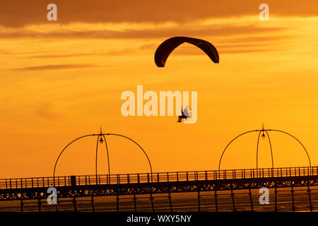 Ein motorbetriebener Handsegler, der in der Abendsonne geschildet wird, nimmt bei leichtem Wind einen motorisierten Flug, wenn die Sonne in Southport untergeht. Paramotor-Flugmaschinen & Stunt-Fliegen bei Sonnenuntergang; eine Handflieger-Silhouette vor orangen Wolken, die mit Propellermaschinen über der irischen Küste fliegen. Stockfoto