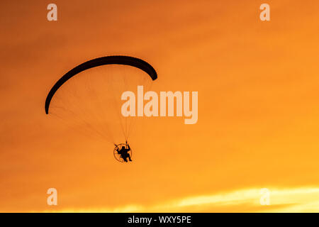 Ein motorbetriebener Handsegler, der in der Abendsonne geschildet wird, nimmt bei leichtem Wind einen motorisierten Flug, wenn die Sonne in Southport untergeht. Paramotor-Flugmaschinen & Stunt-Fliegen bei Sonnenuntergang; eine Handflieger-Silhouette vor orangen Wolken, die mit Propellermaschinen über der irischen Küste fliegen. Stockfoto