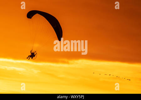 Ein motorbetriebener Handsegler, der in der Abendsonne geschildet wird, nimmt bei leichtem Wind einen motorisierten Flug, wenn die Sonne in Southport untergeht. Paramotor-Flugmaschinen & Stunt-Fliegen bei Sonnenuntergang; eine Handflieger-Silhouette vor orangen Wolken, die mit Propellermaschinen über der irischen Küste fliegen. Stockfoto