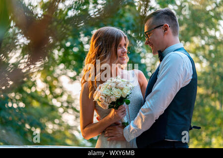Frisch vermählte Paar stehen eng zusammen und umarmen, mit einem Lächeln. Sommer Park mit schönen Sonnenuntergang ist auf Hintergrund und verschwommen. Stockfoto