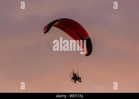 Ein motorbetriebener Handsegler, der in der Abendsonne geschildet wird, nimmt bei leichtem Wind einen motorisierten Flug, wenn die Sonne in Southport untergeht. Paramotor-Flugmaschinen & Stunt-Fliegen bei Sonnenuntergang; eine Handflieger-Silhouette vor orangen Wolken, die mit Propellermaschinen über der irischen Küste fliegen. Stockfoto