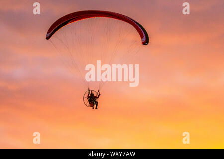 Ein motorbetriebener Handsegler, der in der Abendsonne geschildet wird, nimmt bei leichtem Wind einen motorisierten Flug, wenn die Sonne in Southport untergeht. Paramotor-Flugmaschinen & Stunt-Fliegen bei Sonnenuntergang; eine Handflieger-Silhouette vor orangen Wolken, die mit Propellermaschinen über der irischen Küste fliegen. Stockfoto