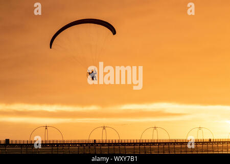 Ein motorbetriebener Handsegler, der in der Abendsonne geschildet wird, nimmt bei leichtem Wind einen motorisierten Flug, wenn die Sonne in Southport untergeht. Paramotor-Flugmaschinen & Stunt-Fliegen bei Sonnenuntergang; eine Handflieger-Silhouette vor orangen Wolken, die mit Propellermaschinen über der irischen Küste fliegen. Stockfoto