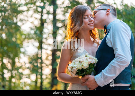 Frisch vermählte Paar stehen eng zusammen und umarmen, mit einem Lächeln. Sommer Park mit schönen Sonnenuntergang ist auf Hintergrund und verschwommen. Stockfoto
