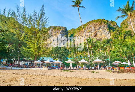 AO NANG, THAILAND - 25 April 2019: Die Linie der Sonnenliegen mit Sonnenschirmen auf den üppigen grünen Garten des Centara Grand Beach, von riesigen Felsen umgeben, auf einem Stockfoto