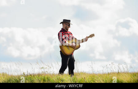 Inspiriert land Musiker. Landhausstil. Sommer Urlaub. Country Music Konzept. Gitarrist Land Sänger stand im Feld Himmel Hintergrund. Wandern Song. United mit der Natur. Stattlicher Mann mit Gitarre. Stockfoto