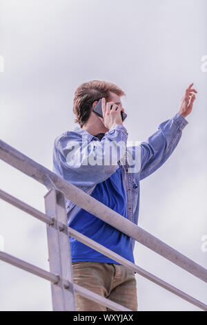 Profil von cute blonde Junge, der in Richtung Himmel, während Sie telefonieren trägt eine blaue Jacke und T-Shirt. Bild vertikal. Stockfoto