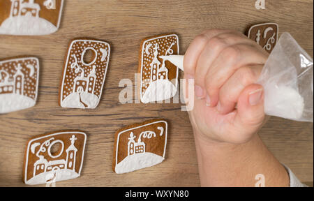 Weibliche hand Detail, wenn Dekoration Weihnachten Lebkuchen durch Vereisung in Spritzbeutel. Gebackene Süßigkeiten mit Weiß lackiert verschneite Kirche auf Holz Hintergrund niedlich. Stockfoto