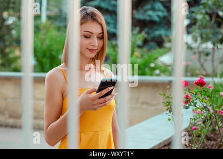 Junge Frau von SMS auf dem Handy betrachten Sie lächelnd auf städtischen Park Hintergrund isoliert. Bild aus dem Büro Jalousien. Mädchen in einem Stockfoto