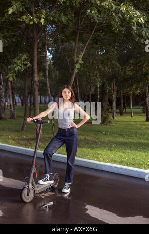 Brunette Mädchen reiten eine Umweltfreundliche elektrische Kick scooter in einem Park bei sonnigem Wetter auf Bürgersteige Stockfoto