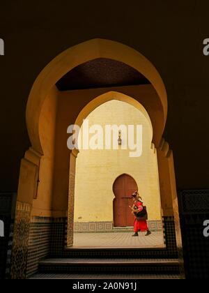 Un portador de agua o 'guerrab' en coloridos trajes tradicionales vistos a través de arcadas del Palacio Moulay Ismail, Meknes, Marruecos Foto de stock