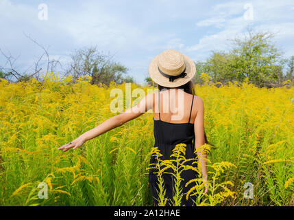 Chica con pelo largo y oscuro en un sombrero entre flores amarillas, vista posterior de una joven mujer Foto de stock
