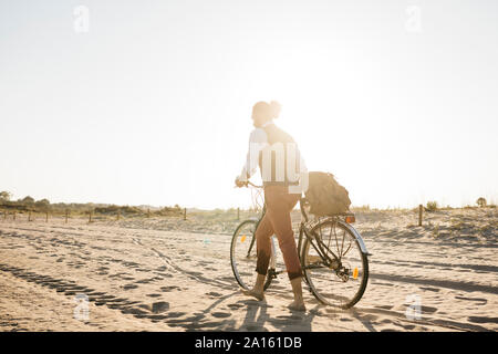 El hombre bien vestido caminando con su bicicleta en una playa en el atardecer.