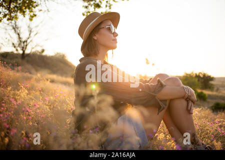Mujer joven llevar sombrero, sentado en la pradera al atardecer