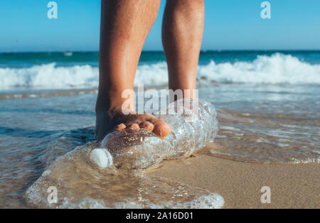 Pie de hombre pisando sobre la botella de plástico vacía para tumbarse en la playa, más cerca Foto de stock