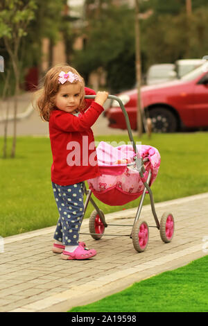 Retrato de un año viejo curly cute little girl cardigan vestidos de rojo y una diadema con un lazo con un juguete rosa paseo en el parque, emulando la polilla Foto de stock
