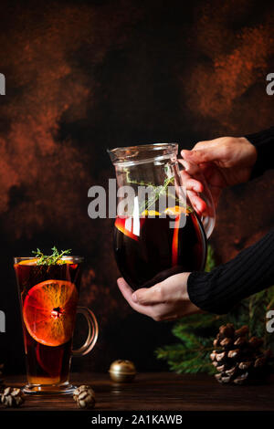 Vino caliente una bebida caliente hecha de vino tinto, cítricos y especias en un vaso sobre una mesa de madera con adornos. Mujeres manos sosteniendo un decantador Foto de stock