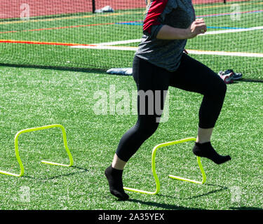 Un atleta de la escuela secundaria femenina se ejecuta sin zapatos sobre amarillo mini banana obstáculos durante la velocidad, fuerza y agilidad formación sobre un césped verde.