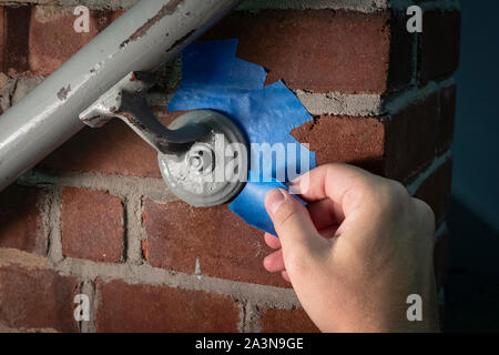 La mano que prepone la barandilla de metal en la pared del ladrillo con la cinta azul del pintor, en preparación para la capa fresca de la pintura Foto de stock