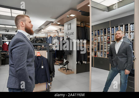 Barbudo cliente masculino vistiendo elegante chaqueta casual mirando en el espejo y elegir ropa en gran boutique. Hombre elegante manteniendo la mano en el guante y posando delante del espejo en la tienda. Concepto de montaje.
