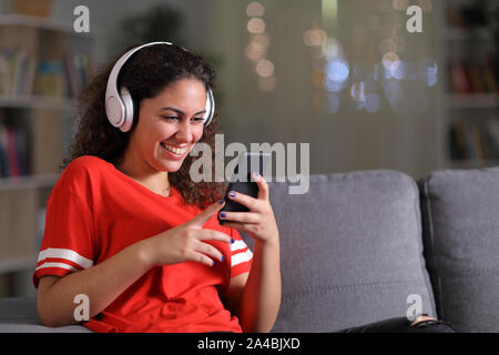 Niña alegre en rojo escuchando música control smart phone sentado en un sofá en el salón en la noche en su domicilio. Foto de stock