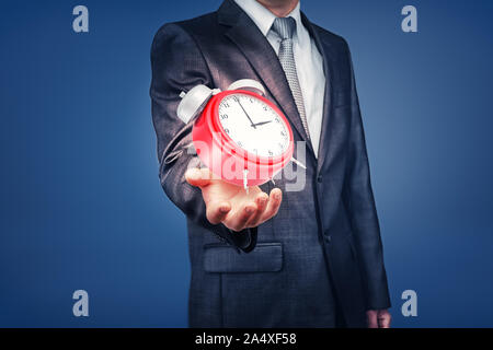 Un reloj de alarma de color rojo brillante se sitúa encima de un brazo abierto del empresario en una visión cercana. Foto de stock