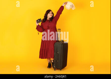 Atractiva mujer viajero del sur de Asia en bata vestido rojo profundo, sombrero planteados en studio sobre fondo amarillo con la maleta con amd antigua cámara de fotos vintage. Foto de stock