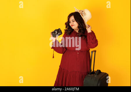 Atractiva mujer viajero del sur de Asia en bata vestido rojo profundo, sombrero planteados en studio sobre fondo amarillo con la maleta con amd antigua cámara de fotos vintage. Foto de stock