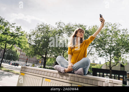 Mujer joven sentado en una caja en la ciudad tomando un selfie