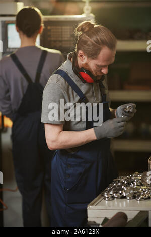 Hombre Barbado en ropa de trabajo y permanente examinar detalles de metal con su colega en el fondo de la planta Foto de stock