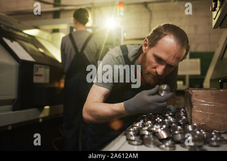 Joven hombre barbado en guantes anticortes examinar detalles de metal mientras trabajaba en la fábrica metalúrgica con la mujer en el fondo Foto de stock