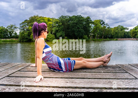Modelo bastante joven con hermoso cabello púrpura se asienta en un embarcadero de madera mirando al lago teniendo en la vista, vestido a rayas y zapatos planos