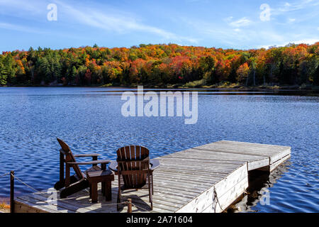 Dos sillas Muskoka sentado en un muelle de madera frente a un lago en una tranquila temporada de otoño en un día soleado Foto de stock
