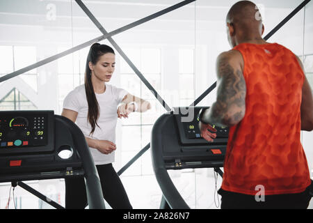 Entrenador atento mirando fitness tracker mientras está de pie cerca de deportista afroamericano corriendo en cinta