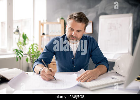 Arquitecto maduro serio dibujo nuevo boceto en papel mientras está sentado al escritorio
