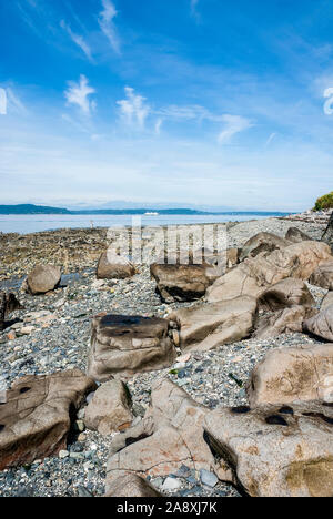 Alki playa rocosa con una mujer solitaria en la orilla y un ferry en el Canal Puget en el oeste de la ciudad de Seattle, Washington. Foto de stock