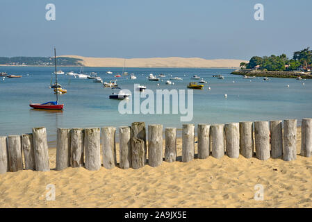 Playa con troncos de madera proteger las dunas de Cap-Ferret, barcos y la duna de Pilat en el fondo. Comuna de la Gironde en Francia Foto de stock
