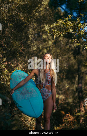 Joven Mujer rubia con un surfboard caminando en el bosque, mirando hacia arriba Foto de stock
