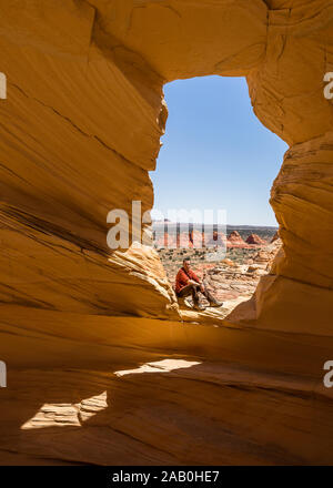 Hombre sentado mirando a la cámara en un arco desde el alcove Teepee, mirando a lo largo de formaciones de arenisca del Coyote Buttes north wilderne