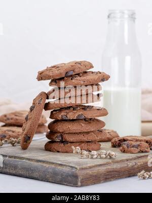 Pila de galletas con trocitos de chocolate redondo sobre una tabla de madera marrón, detrás de una botella de leche.