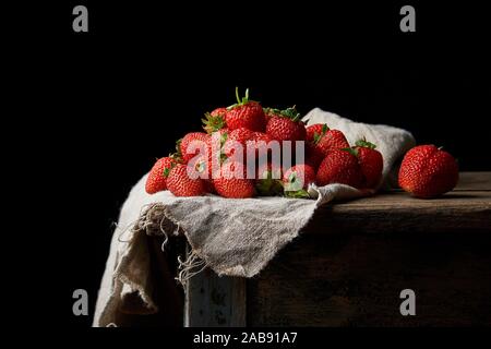 Montón de frescas fresas rojas maduras en una servilleta de lino gris, mesa de madera, fondo negro.