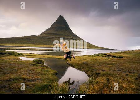 Excursionista joven salta sobre un arroyo en la montaña Kirkjufell en Islandia Foto de stock