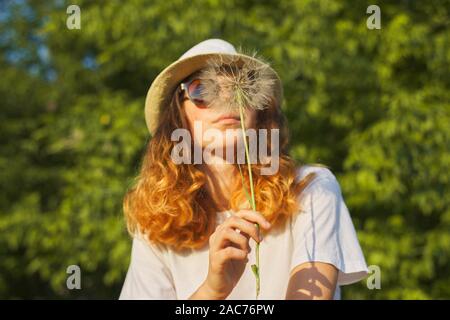 De verano al aire libre retrato de una adolescente romántica en el sombrero con grandes y esponjosas jaramago en la naturaleza, soplando sobre el diente de león en seco
