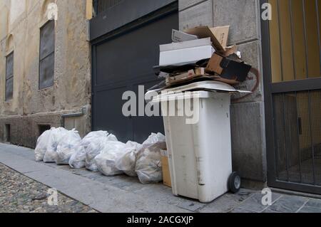 Vista de un recipiente de plástico blanco con exceso de desechos de cartón y muchas bolsas de basura blanca recostada sobre la calle de Padua en Italia Foto de stock