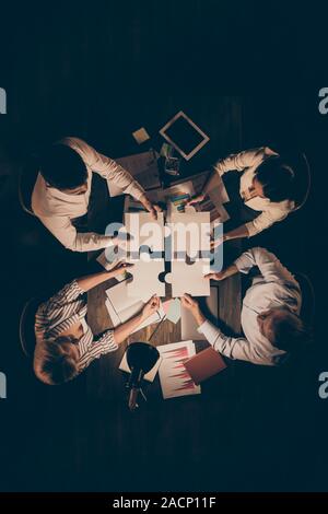 Ángulo alto arriba ver foto vertical de cuatro empresarios colegas sentados círculo mesa trabajando tarde noche sujetando las piezas del rompecabezas de papel encontrar la unidad