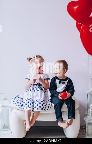 Niña y Niño sentado en una silla blanca cerca de globos con forma de corazón. Chica lamiendo un círculo rojo. Día de San Valentín concepto