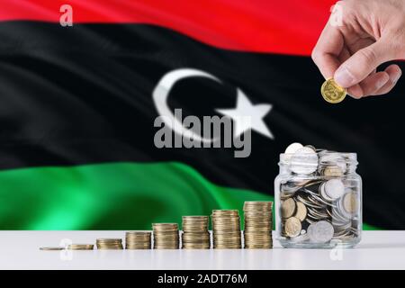 Hombre de negocios mantiene monedas poniendo en vidrio, Libia ondear la bandera en el fondo. Concepto de negocio y finanzas. Ahorro de dinero. Foto de stock