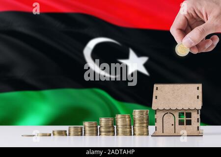 Hombre sujetando monedas poniendo en casa de madera hucha, Libia ondear la bandera en el fondo. Ahorro de dinero para la hipoteca. Foto de stock