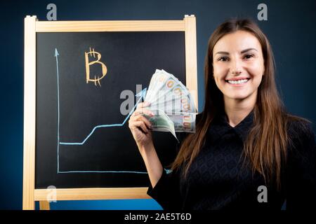 Gráfico de crecimiento Bitcoin se dibuja en una pizarra negra, una mujer sonriente está de pie cerca. El mercado financiero de divisas Foto de stock